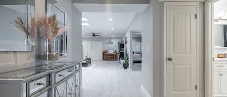 A modern kitchen with a central island, a dining area with a chandelier, and a hallway leading to a bedroom.