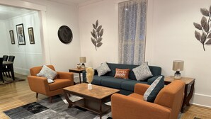 A living room with a sofa, two armchairs, a coffee table, and a rug. There are wall decorations and a lamp on a side table.