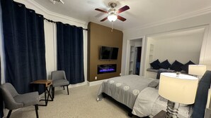 A hotel room with a bed, a TV mounted on the wall, a ceiling fan, and a small table with a lamp.