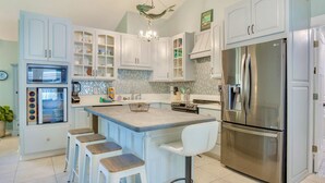 Una cocina con armarios blancos, un refrigerador de acero inoxidable, un microondas y una estufa. Hay taburetes de bar y una pequeña isla con un lavamanos.