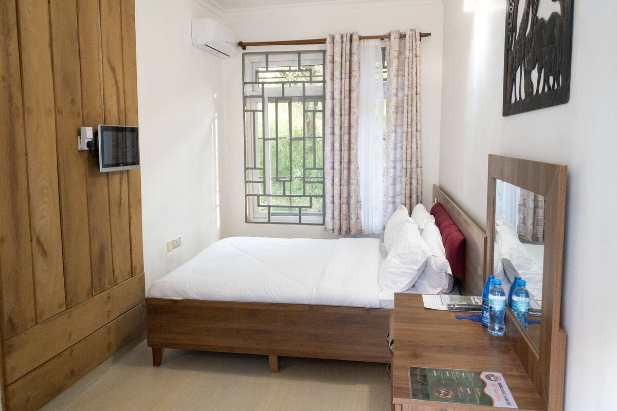 A bedroom with a wooden bed, a window with curtains, a bedside table with water bottles, and a wall-mounted air conditioner.