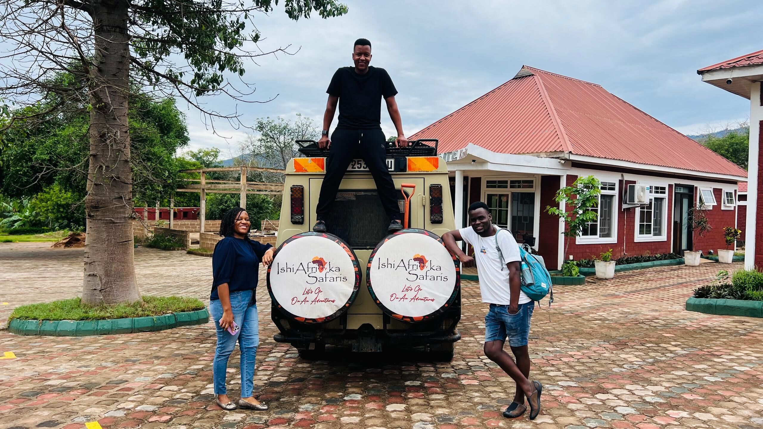A building with a red roof and white walls, a paved area with a tree, and a vehicle with two circular signs reading "Ishi Africa Safaris".