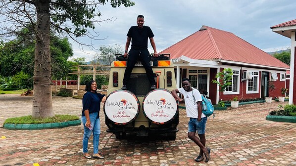 A building with a red roof and white walls, a paved area with a tree, and a vehicle with two circular signs reading "Ishi Africa Safaris".