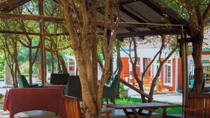 An outdoor seating area with wooden tables and chairs, surrounded by trees and a covered structure. - Mkomazi Hotels and Campsite (Kisiwani)