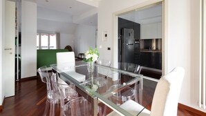A modern dining area with a glass table and clear chairs, a green accent wall, and a kitchen in the background.