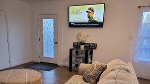 A living room with a sofa, a coffee table, a TV mounted on the wall, a door with a frosted glass panel, and a small side table with a sign.
