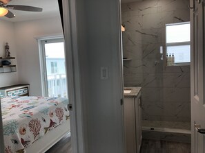 Una camera da letto con un letto, un comodino e una ventola a soffitto. C'è un bagno con una doccia, un lavandino e una finestra.