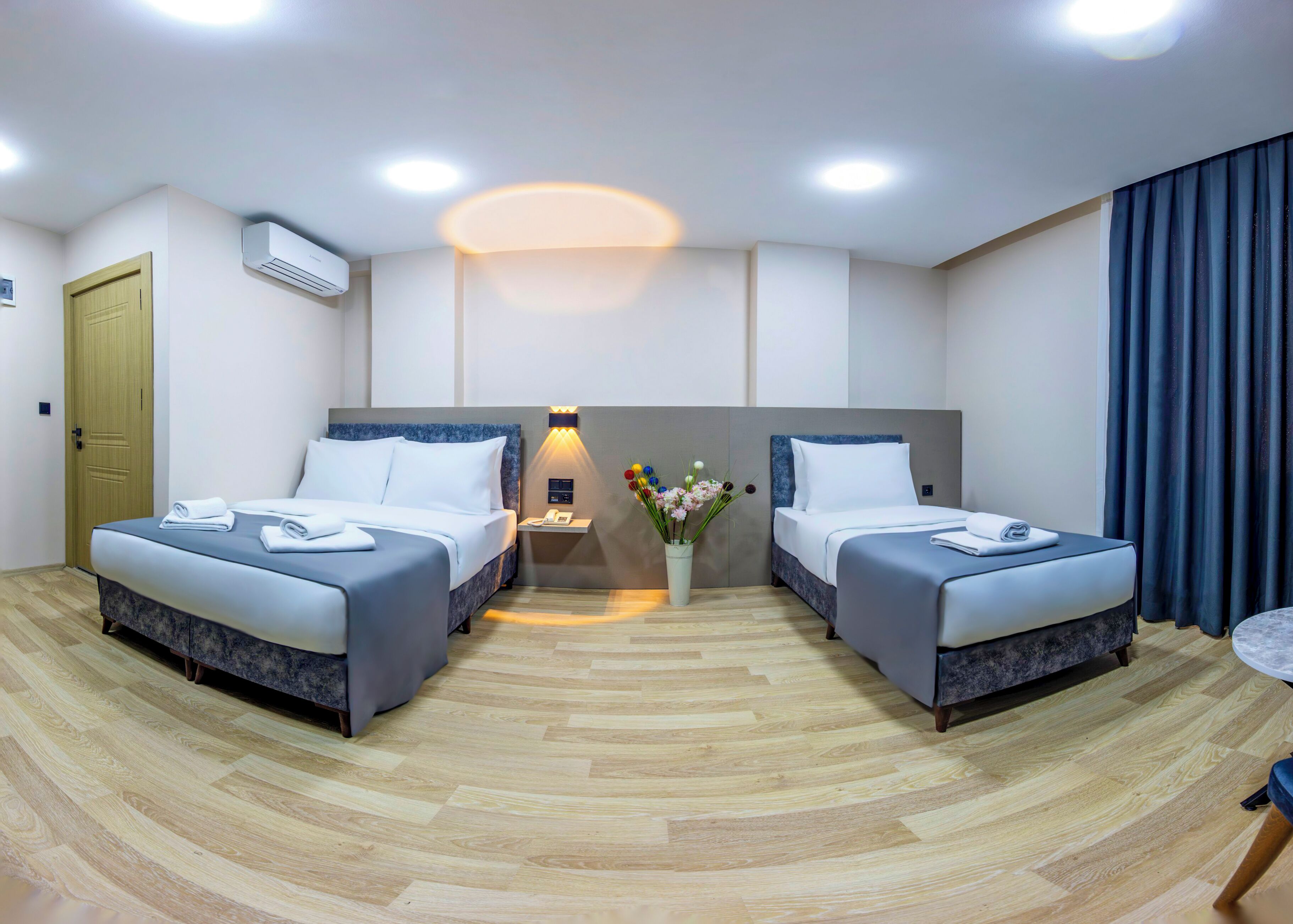 A hotel room with two beds, a bedside table with a lamp, a vase with flowers, and a wooden floor.