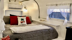 A cozy bedroom with a bed, a bedside table, a window with curtains, and a wall-mounted air conditioner. - Hocking Hills Airstream & Outdoor Living (McArthur)