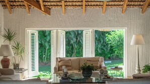 Une pièce avec un canapé, une table basse en verre et une vue sur un jardin luxuriant, visible à travers des volets ouverts.