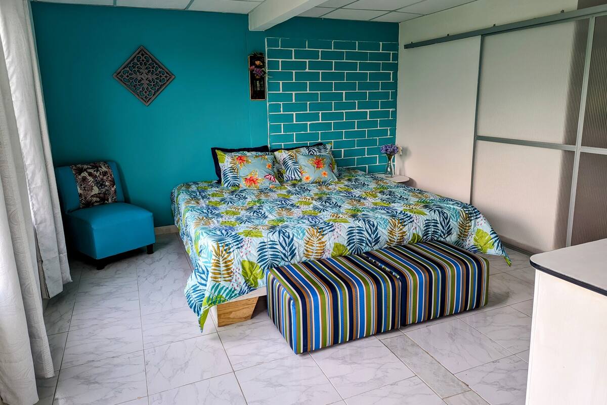 Une chambre avec un mur de couleur turquoise, une tête de lit à motifs de briques, une couvre-lit à fleurs, un banc à rayures et une petite table d’appoint avec un vase.