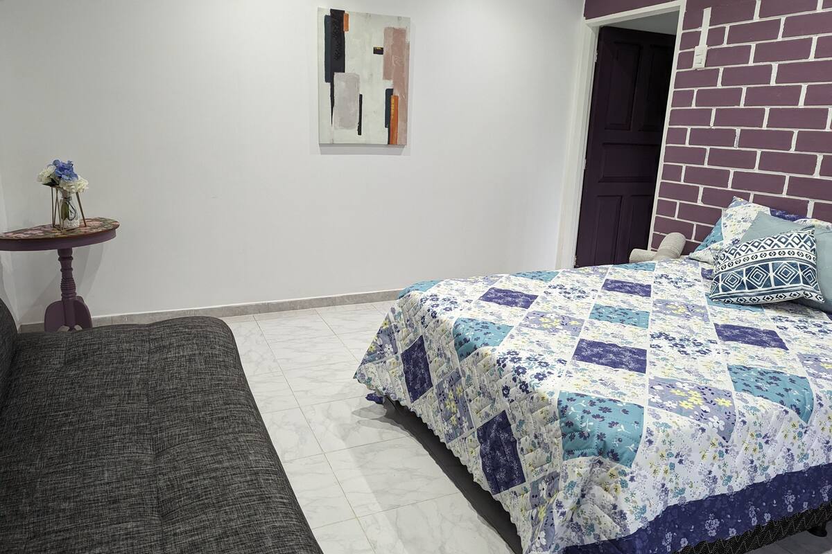 Une chambre à coucher avec un lit, une petite table sur laquelle est posé un vase, et un mur orné d’un tableau.