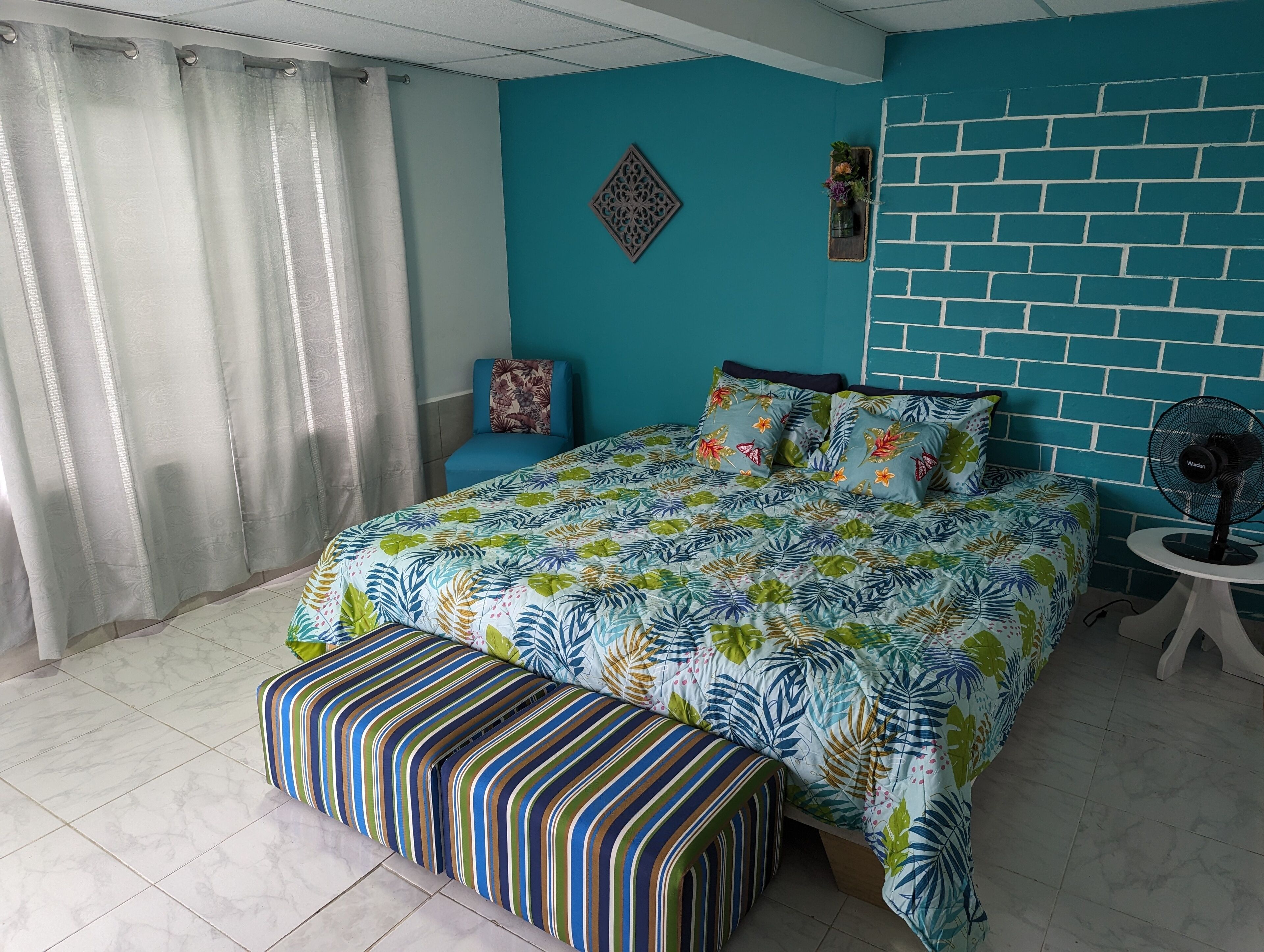 A bedroom with a bed, a bench, a chair, a wall-mounted fan, and a decorative wall piece.