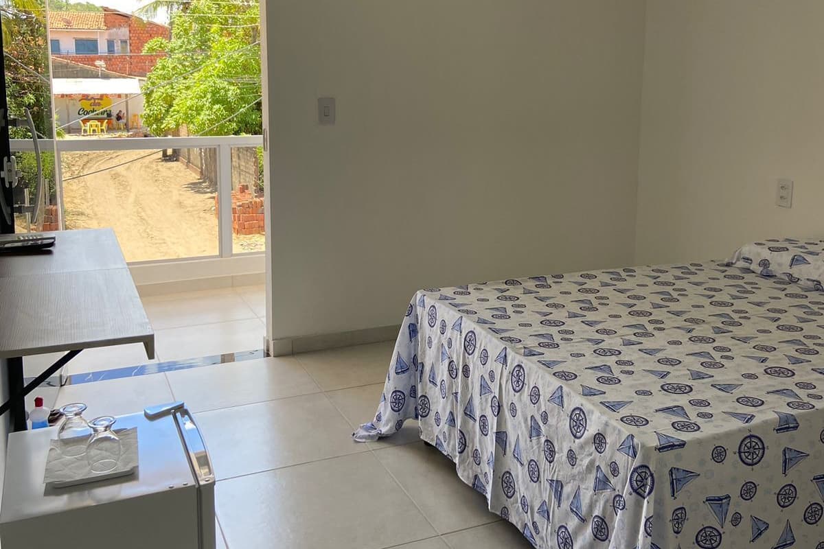 Ein Zimmer mit einem Bett, einem Tisch und einem Stuhl. Ein Fenster bietet Blick auf Gebäude und Bäume.