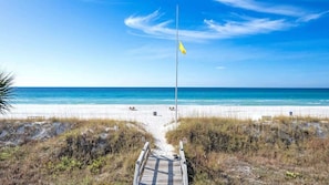 Une plage avec un mât de drapeau, une promenade en bois menant à la plage et un ciel bleu dégagé.