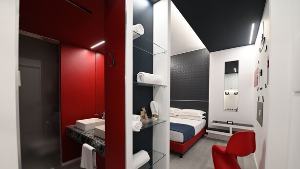 Camera d'albergo moderna con una parete rossa a contrasto, box doccia in vetro, un letto bianco e una poltrona rossa.