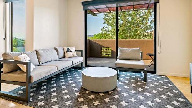Un salon moderne comprenant un canapé, un fauteuil et un pouf, un tapis à motifs et une porte-fenêtre coulissante donnant sur un espace extérieur.