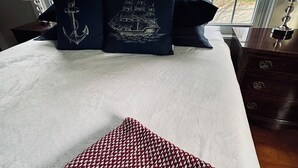 A bed with navy blue pillows featuring a ship and anchor design, a red and white houndstooth throw, and a view of trees through large windows. - Captain's 2BR. Authenticity. Value, Near everything. Yard. Treehouse. No fees.  (Bath)