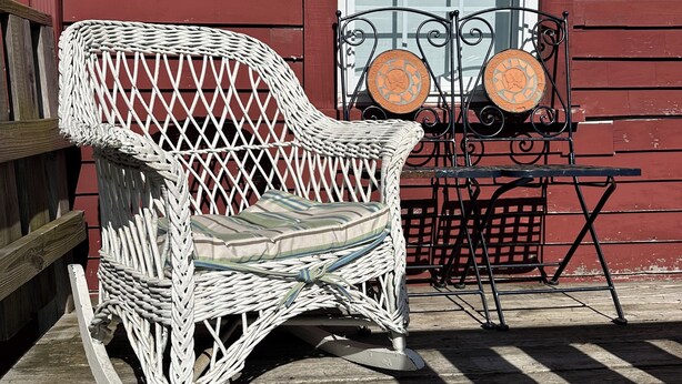 A rocking chair and a small table with a decorative plate, both placed on a wooden deck in front of a red building with a white door. - Captain's 2BR. Authenticity. Value, Near everything. Yard. Treehouse. No fees.  (Bath)
