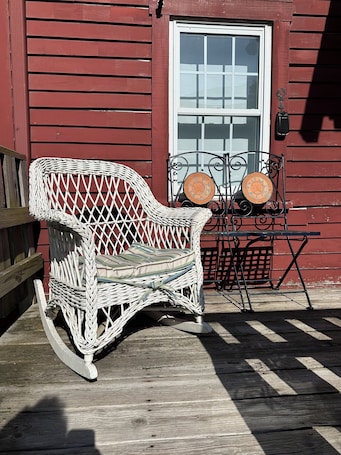 A rocking chair and a small table with a decorative plate, both placed on a wooden deck in front of a red building with a white door.