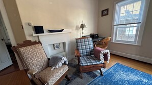 A cozy living room with a fireplace, two armchairs, a blue rug, and a window with blinds. - Captain's 2BR. Authenticity. Value, Near everything. Yard. Treehouse. No fees.  (Bath)