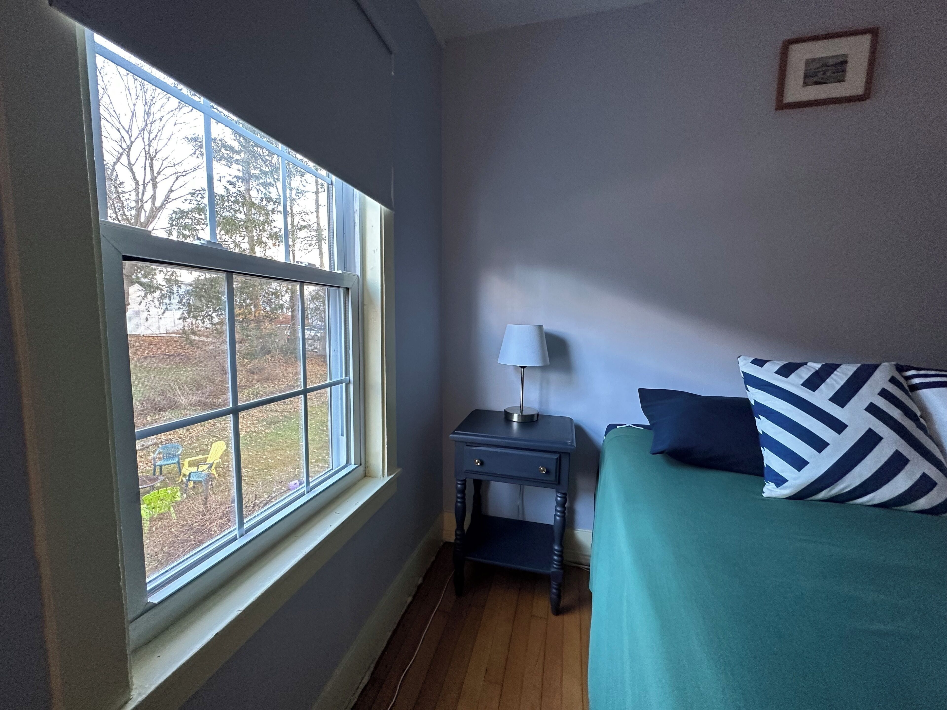 Una camera da letto con una grande finestra, un comodino con una lampada, un letto con copriletto verde e cuscini a righe, e un quadro appeso al muro.