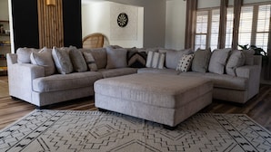 A modern living room with a sectional sofa, ottoman, patterned rug, and a dartboard on the wall.