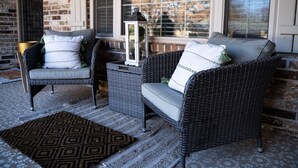 A wicker chair and sofa set with cushions, a lantern, and a patterned rug on a porch with a brick wall and windows.