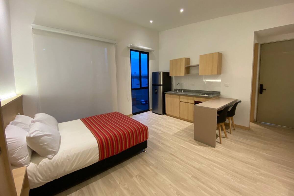 A modern hotel room with a bed, a desk, and a kitchenette.