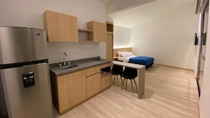 A compact kitchen with a refrigerator, sink, and wooden cabinets. A bed with a blue mattress is visible in the background. - Hotel Estar (Manizales)