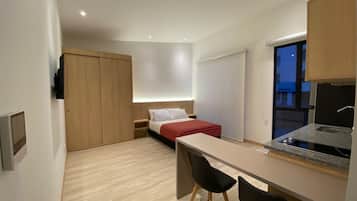 Una habitación compacta con cama, escritorio, dos sillas, una kitchenette pequeña y un armario con puerta corrediza.