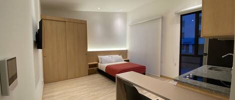 Una habitación compacta con cama, escritorio, dos sillas, una kitchenette pequeña y un armario con puerta corrediza.