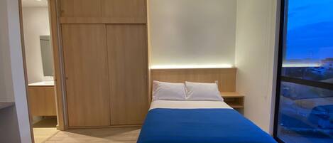 Un dormitorio con cama, armario, mesita de noche y un amplio ventanal con vistas.