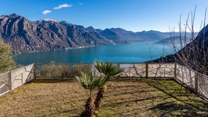 A view of a mountainous landscape with a lake, a fenced area with a palm tree, and a clear sky. - Xino Terrace View by Interhome (Fonteno)