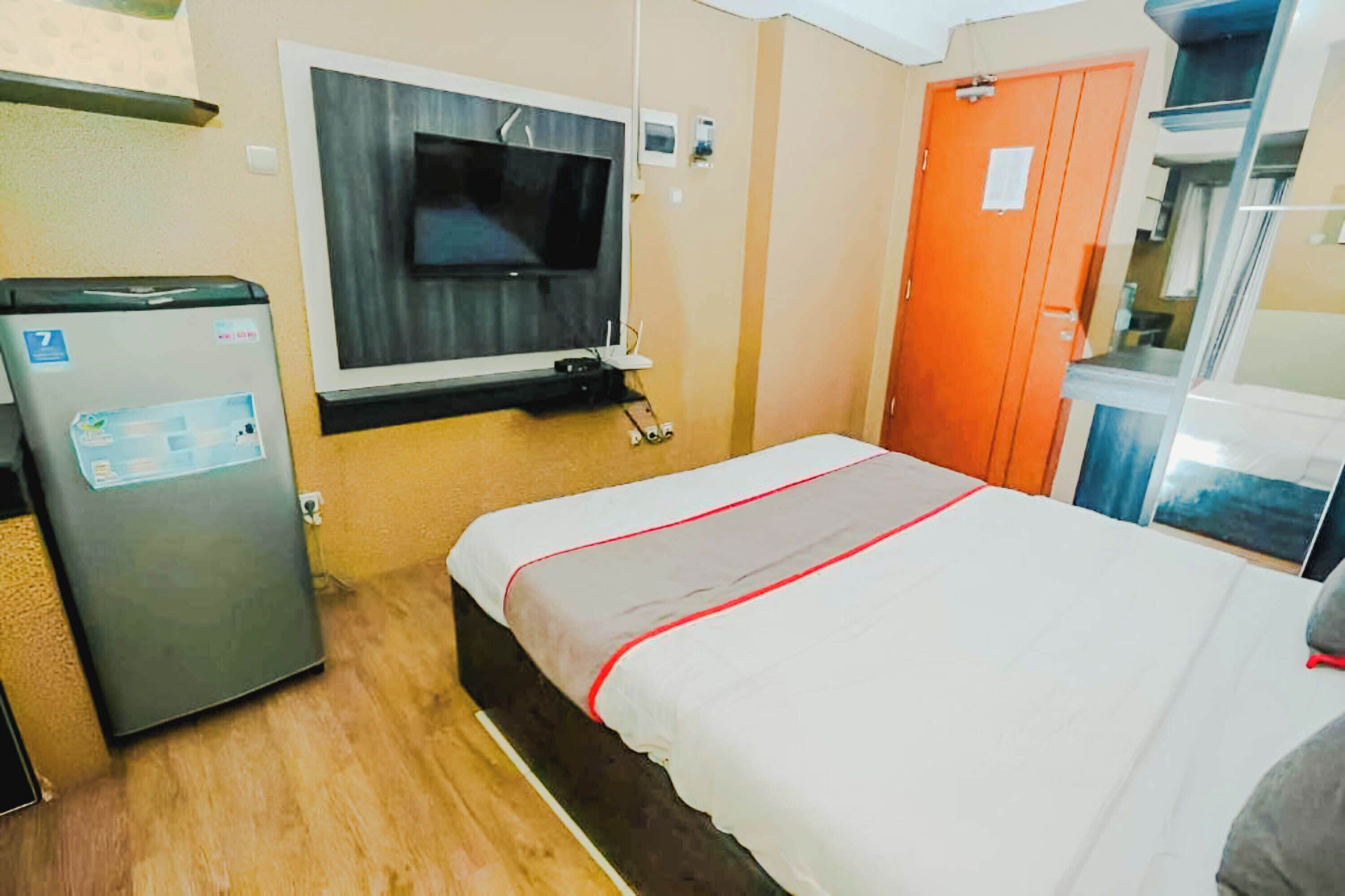 Hotellihuone, jossa on sänky, seinälle asennettu televisio, jääkaappi ja oranssi ovi.