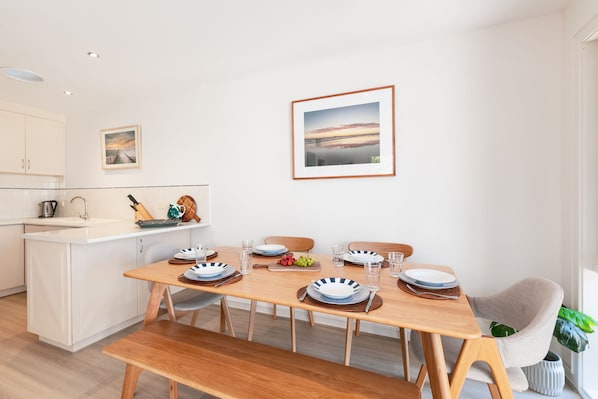 A modern dining area with a wooden table set for four, white cabinetry, and a kitchen in the background. - Lookout 100m to the Ocean (Ocean Grove)