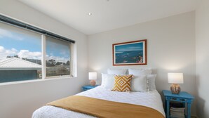 Une chambre à coucher avec un grand lit, deux tables de chevet, une fenêtre donnant sur des immeubles et un tableau encadré au mur.