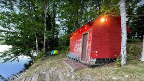 Una cabaña roja pequeña con una terraza de madera y un banco, ubicada cerca de un cuerpo de agua rodeado de árboles.