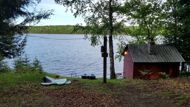 Una cabaña junto al lago, con fachada roja y techo de metal, rodeada de árboles y un muelle con barcos.