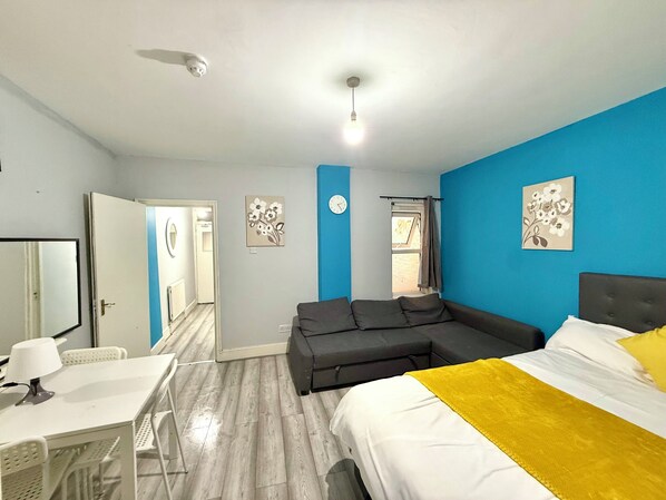 A hotel room with a bed, a sofa, a desk, and a chair. There is a window with curtains, a clock on the wall, and a ceiling light.