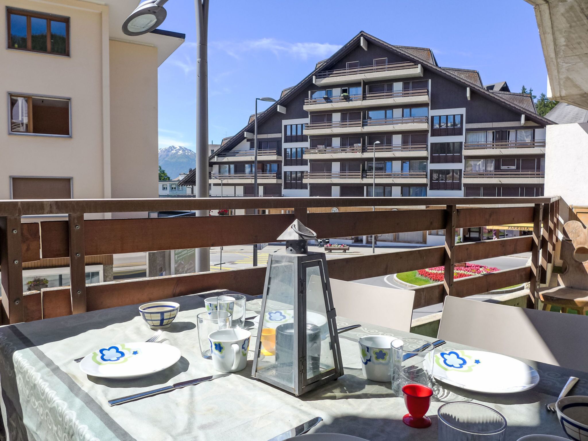 A dining table set with plates, cutlery, and glasses, overlooking a building with multiple balconies and a mountain in the background.