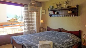 Une chambre à coucher avec un grand lit, un luminaire suspendu, une fenêtre avec des rideaux et une étagère contenant des livres et des objets décoratifs.