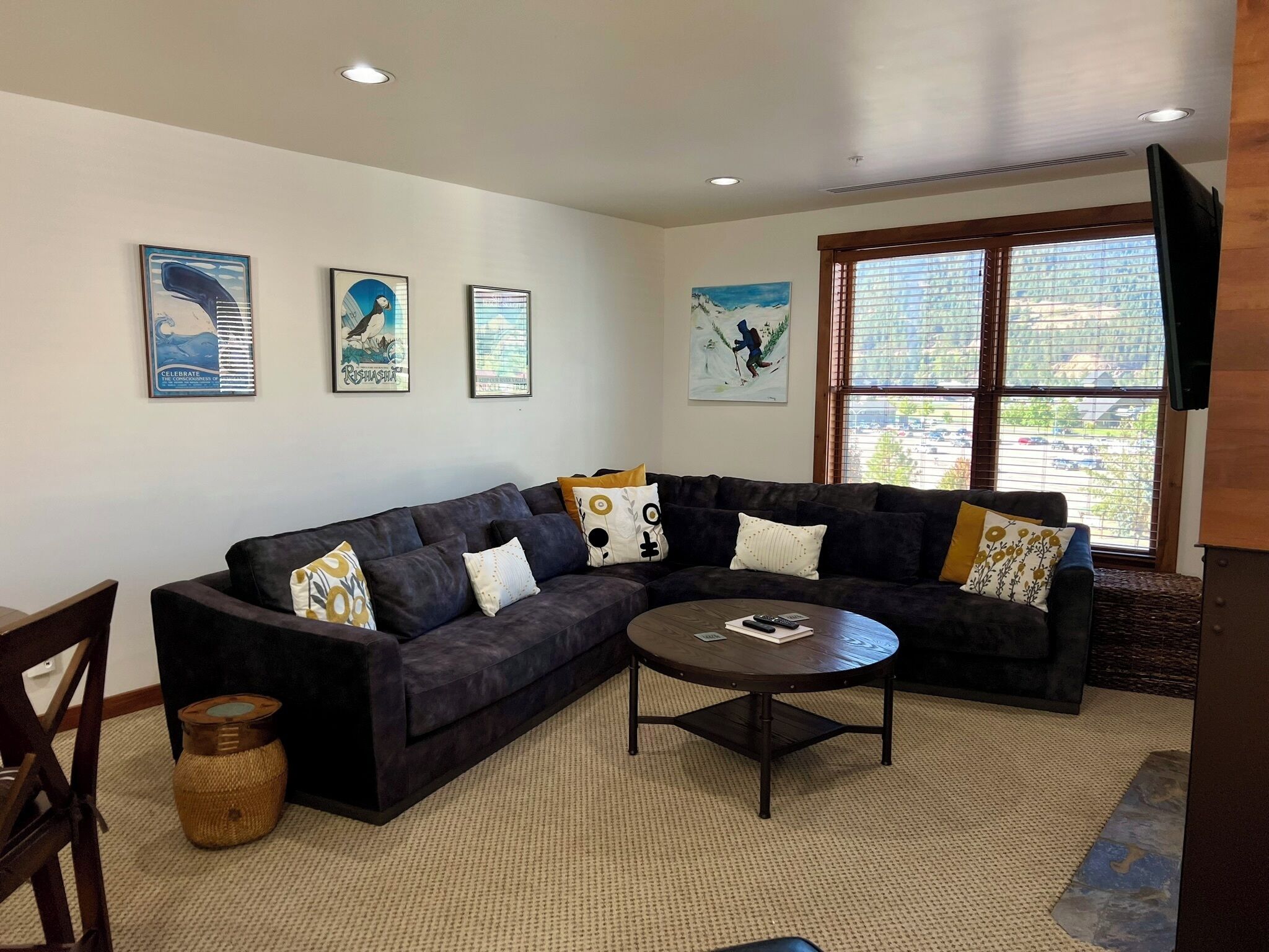 A living room with a dark-colored sectional sofa, a round coffee table, and a large window with blinds.