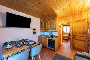 A wooden interior with a dining table, chairs, a kitchen area, and a bedroom visible through an open door.