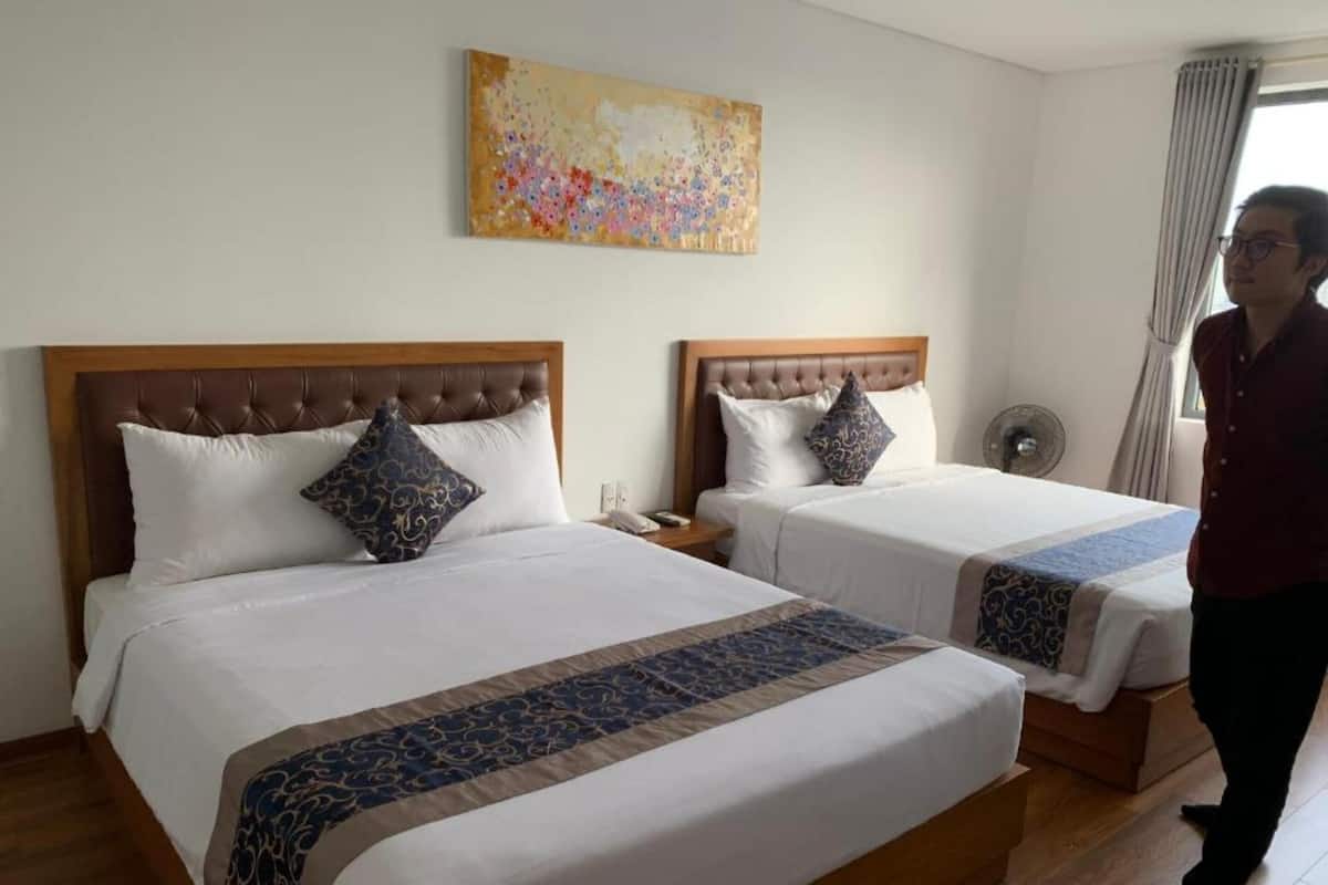 A hotel room with two beds, a wooden headboard, a decorative pillow, a bedside table, a phone, and a painting on the wall.