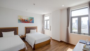 A hotel room with two beds, a painting on the wall, a window with a city view, and a desk with a water bottle. - Sunset Sea Hotel (Da Nang)
