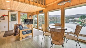 A cozy cabin with a wooden interior, a large window overlooking a lake, a television, and wicker furniture.