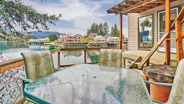 A glass table and chairs on a deck with a wooden railing overlooking a body of water and a residential area.