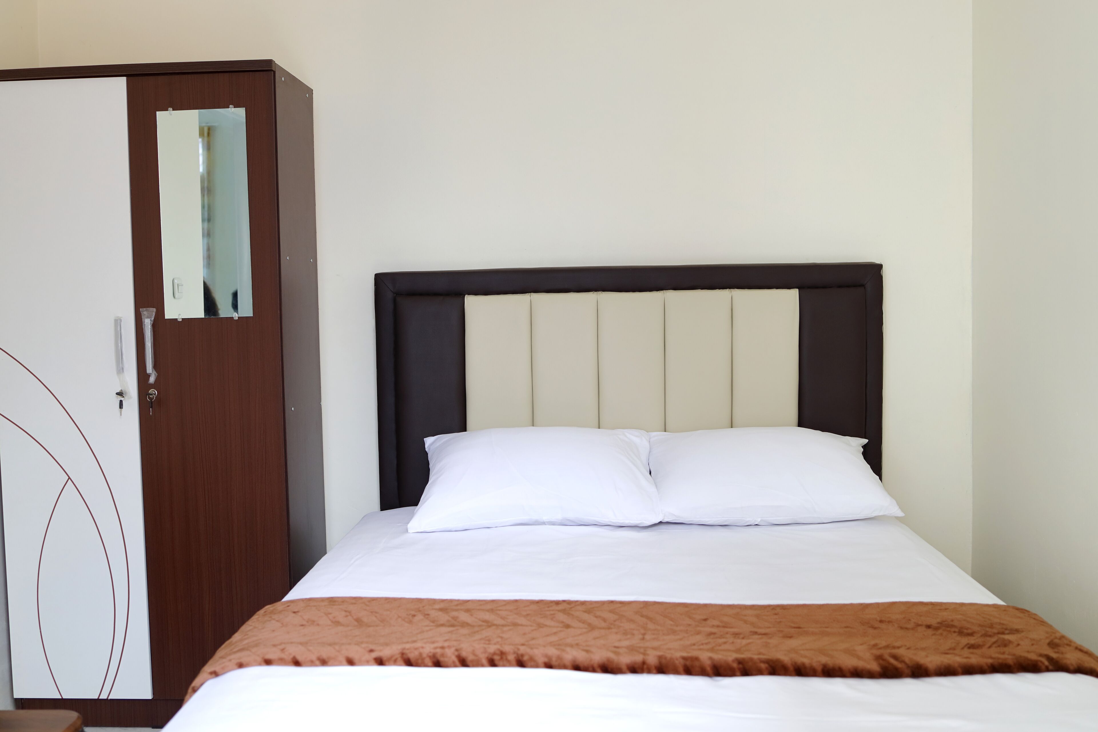A neatly made bed with a brown throw blanket, a wooden wardrobe with a mirror, and a plain wall.