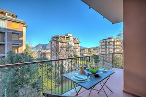 Varanda com mesa posta para refeições, com vista para uma paisagem montanhosa e edifícios vizinhos.